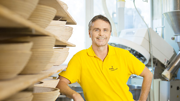 ein Mann in gelbem T-Shirt mit dem ezeb-Logo lehnt sich an ein großes Regal, das voller Brotkörbe ist; im Hintergrund sieht man einen industriellen Mixer